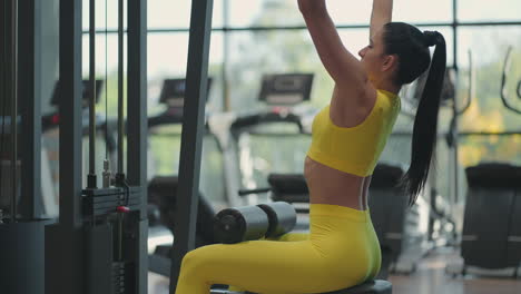 A-young-brunette-woman-in-a-yellow-suit-is-doing-exercises-for-the-muscles-of-the-back-on-the-simulator.-Hispanic-woman-pulls-a-rope-on-the-simulator.-Simulated-pull-ups